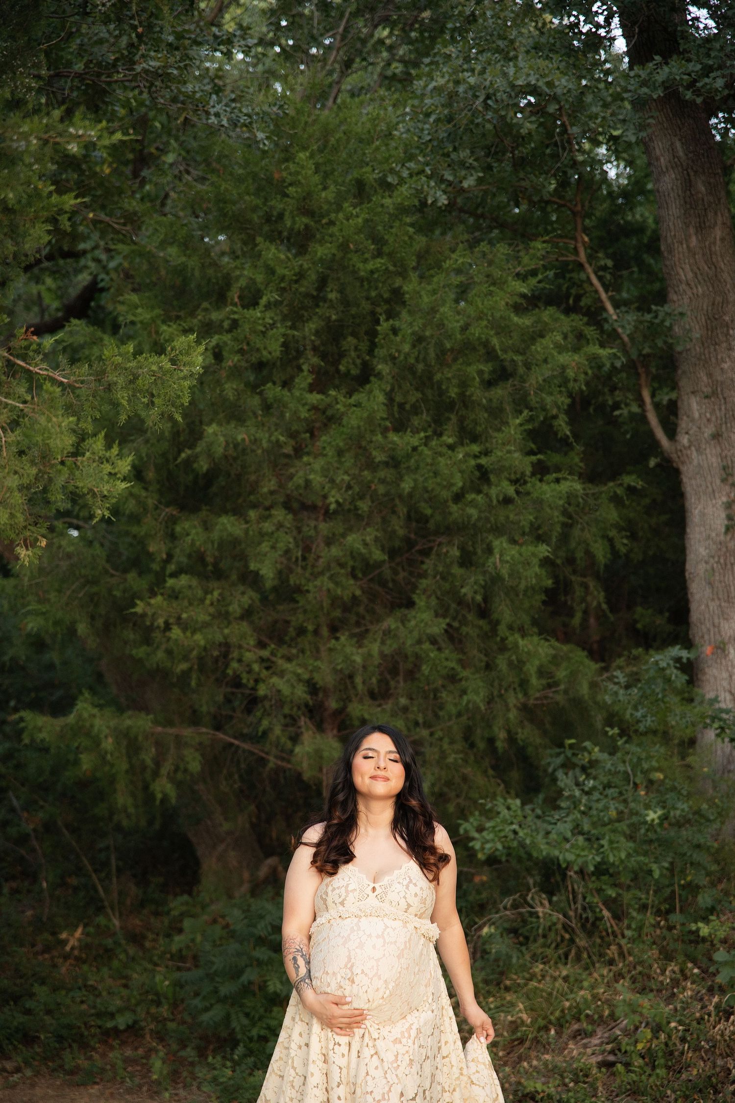 Pregnant woman in cream lace dress standing outdoors among lush green trees, cradling her baby bump with eyes closed.