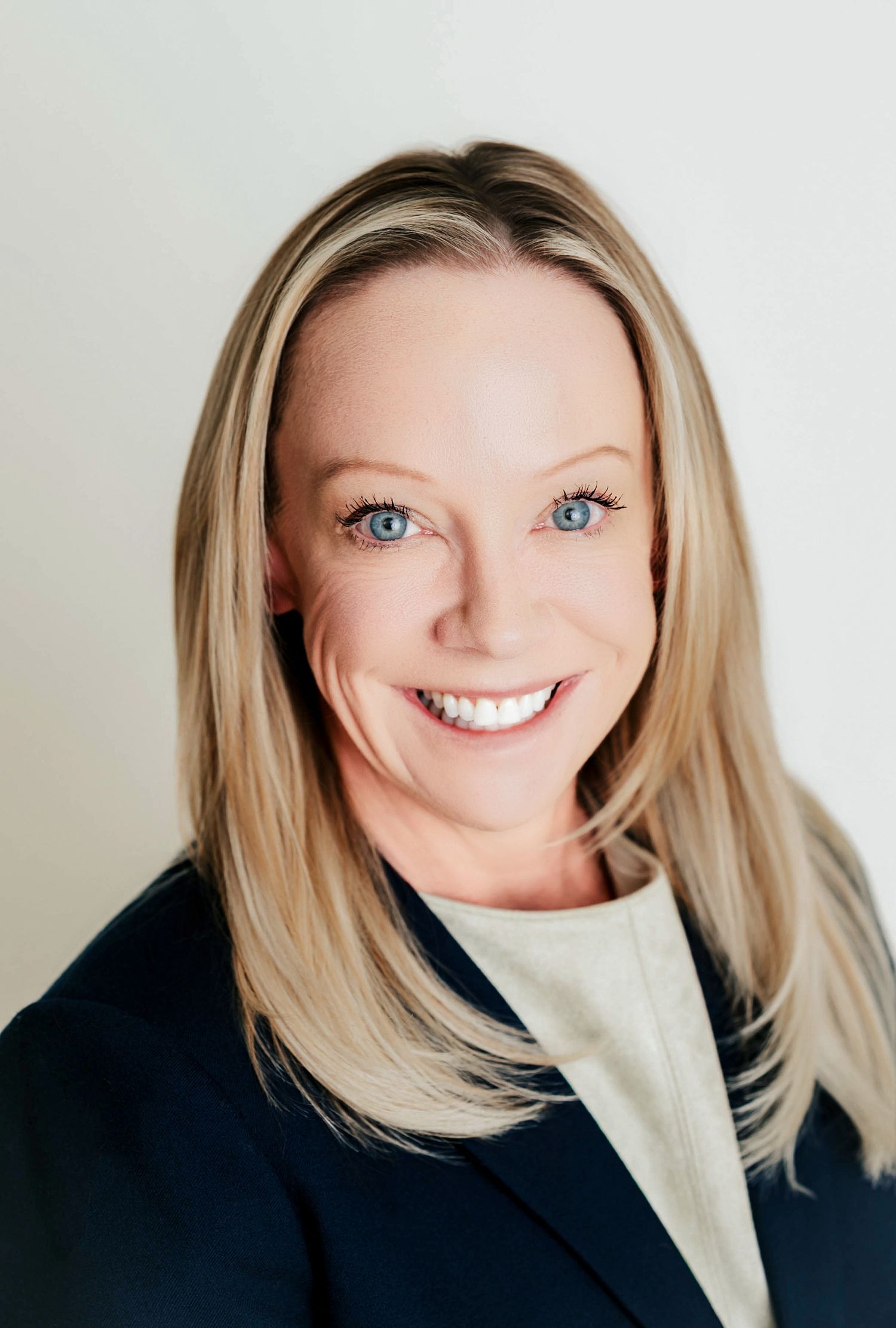 Professional headshot of a person with blonde hair wearing a navy blazer and light colored top against a white background.