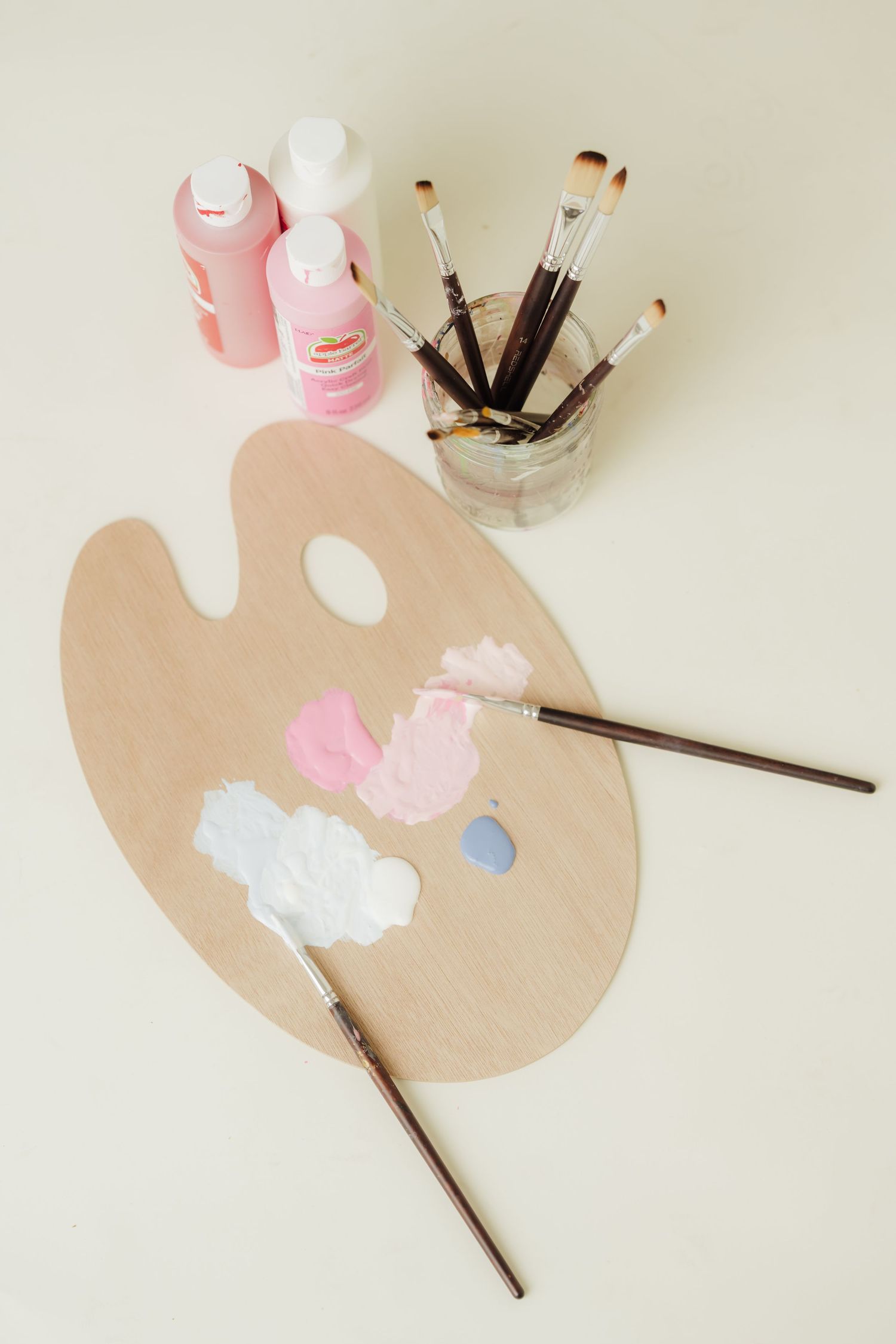 Wooden artist palette with pink and white paint, brushes, and paint bottles on a light background.