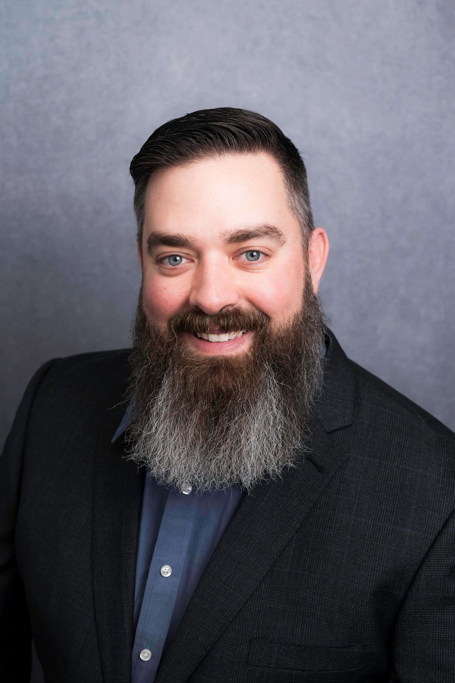 Professional headshot of someone wearing a dark suit jacket and blue shirt with a full grey beard against grey background.