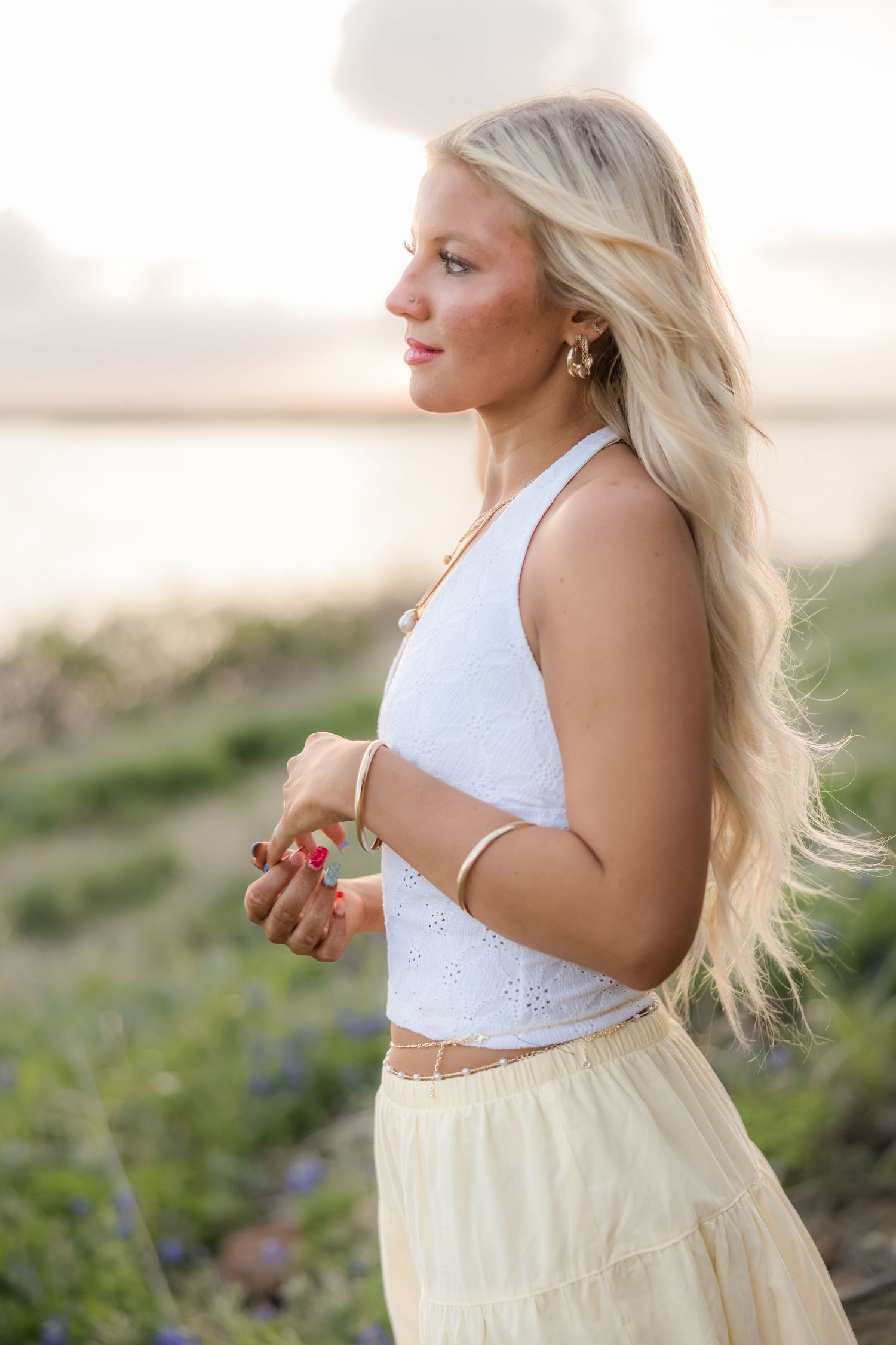 Blonde woman in white crop top and yellow skirt standing outdoors at sunset near a field of wildflowers.