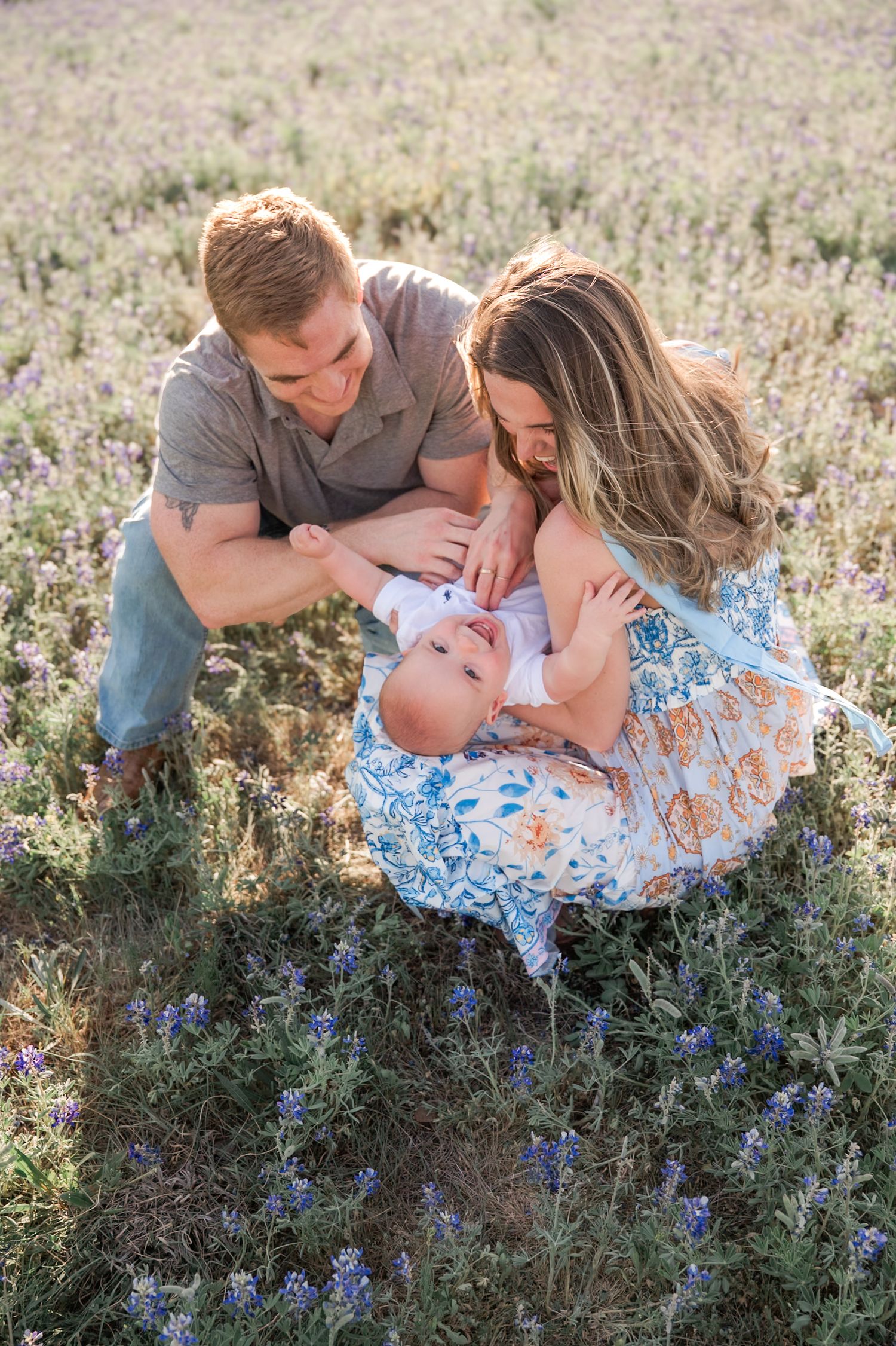 A young family sits in a blooming bluebonnets  field, parents lovingly gazing at their newborn baby outdoors.Northlake Tx, Knot & Wing Photo