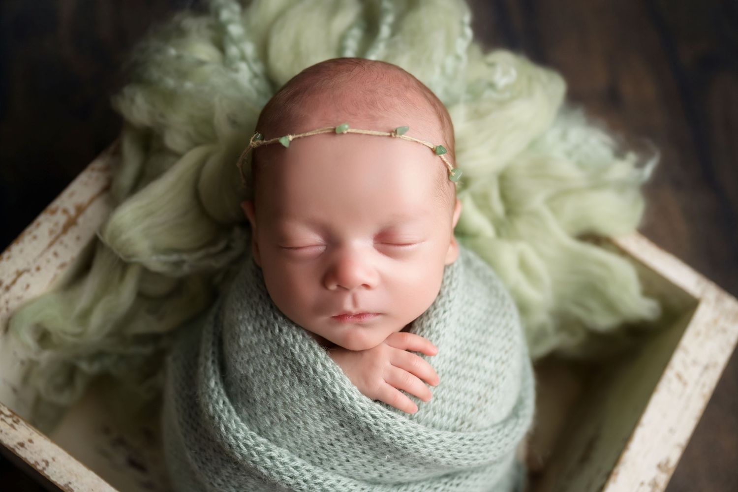 Sleeping newborn baby swaddled in soft green wrap with floral headband resting in white wooden box.