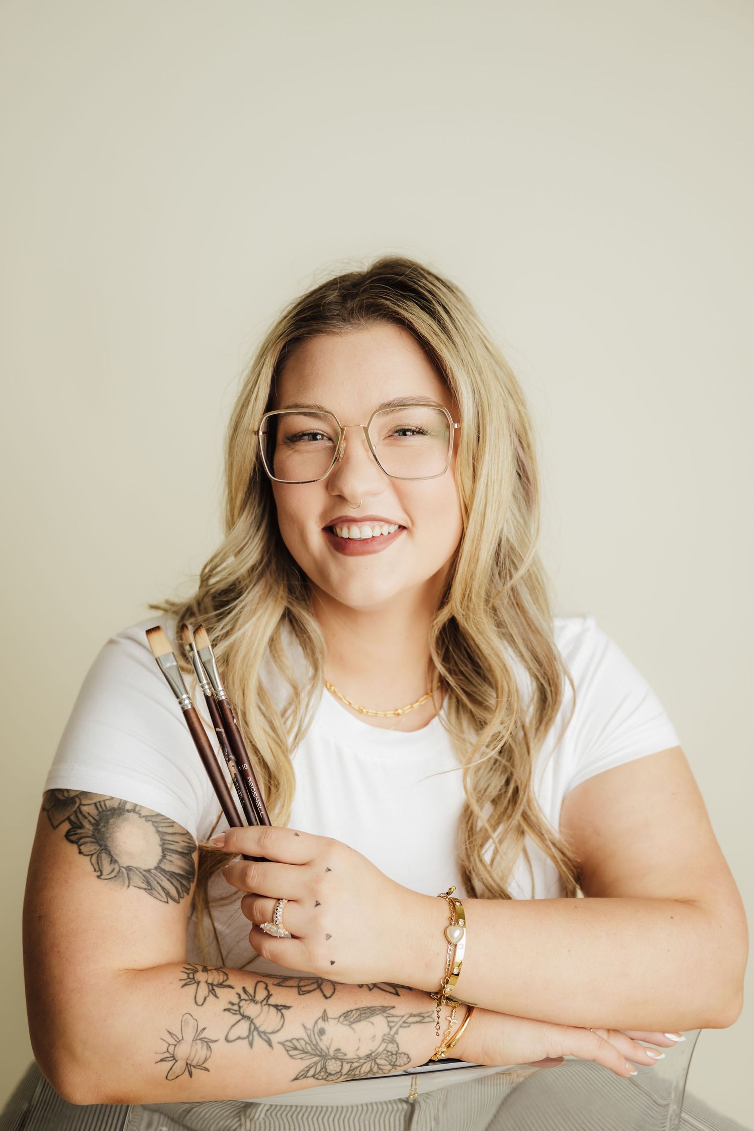 Smiling blonde artist with glasses and tattoos holding paintbrushes, wearing a white t-shirt and gold jewelry.