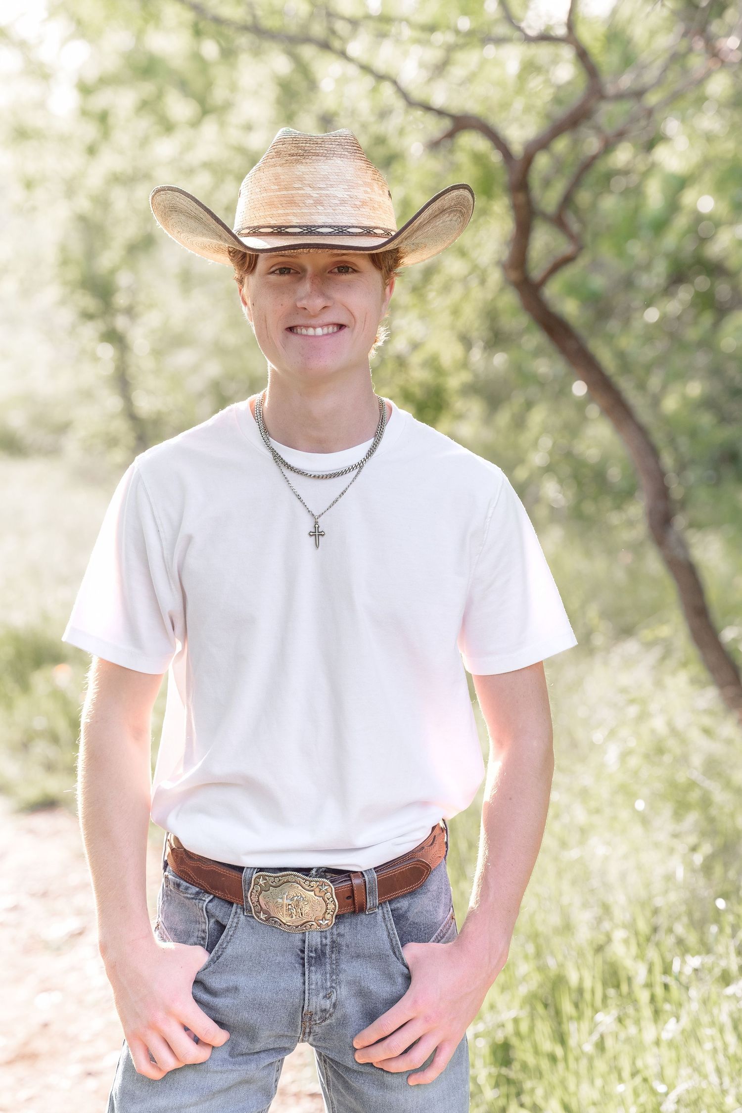 Smiling young man in straw cowboy hat, white t-shirt, jeans, and cross necklace standing outdoors.