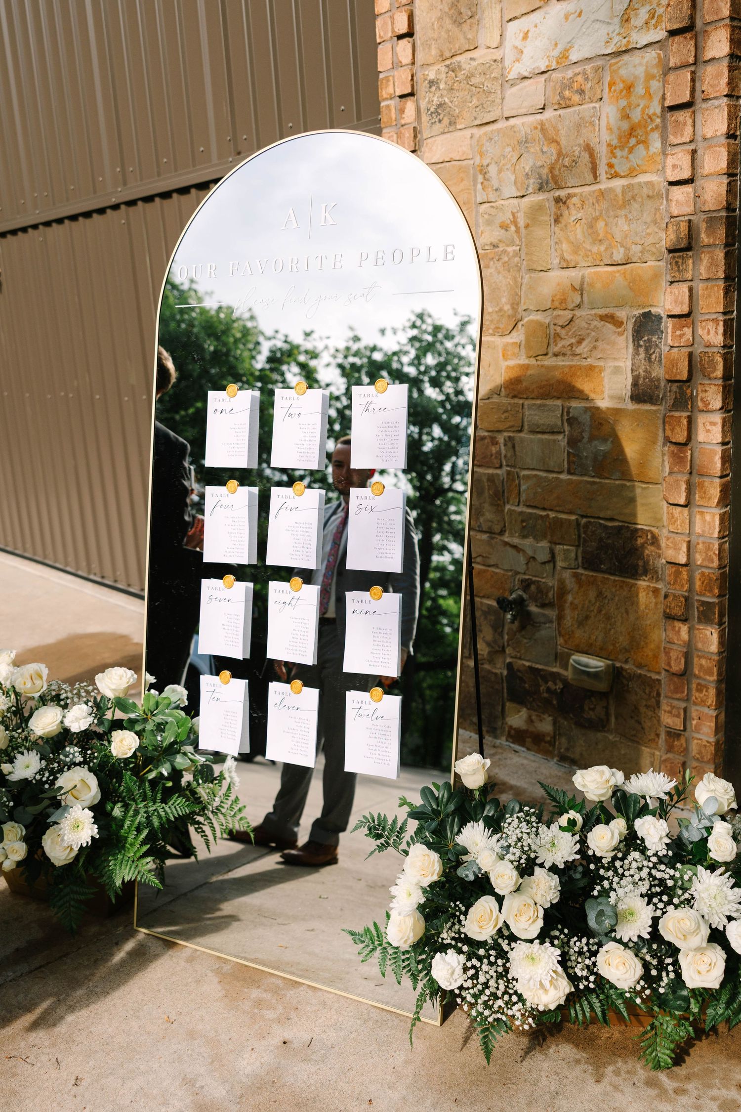 Arched mirror wedding seating chart with hanging cards, framed by white rose and greenery floral arrangements beside a rustic stone wall.