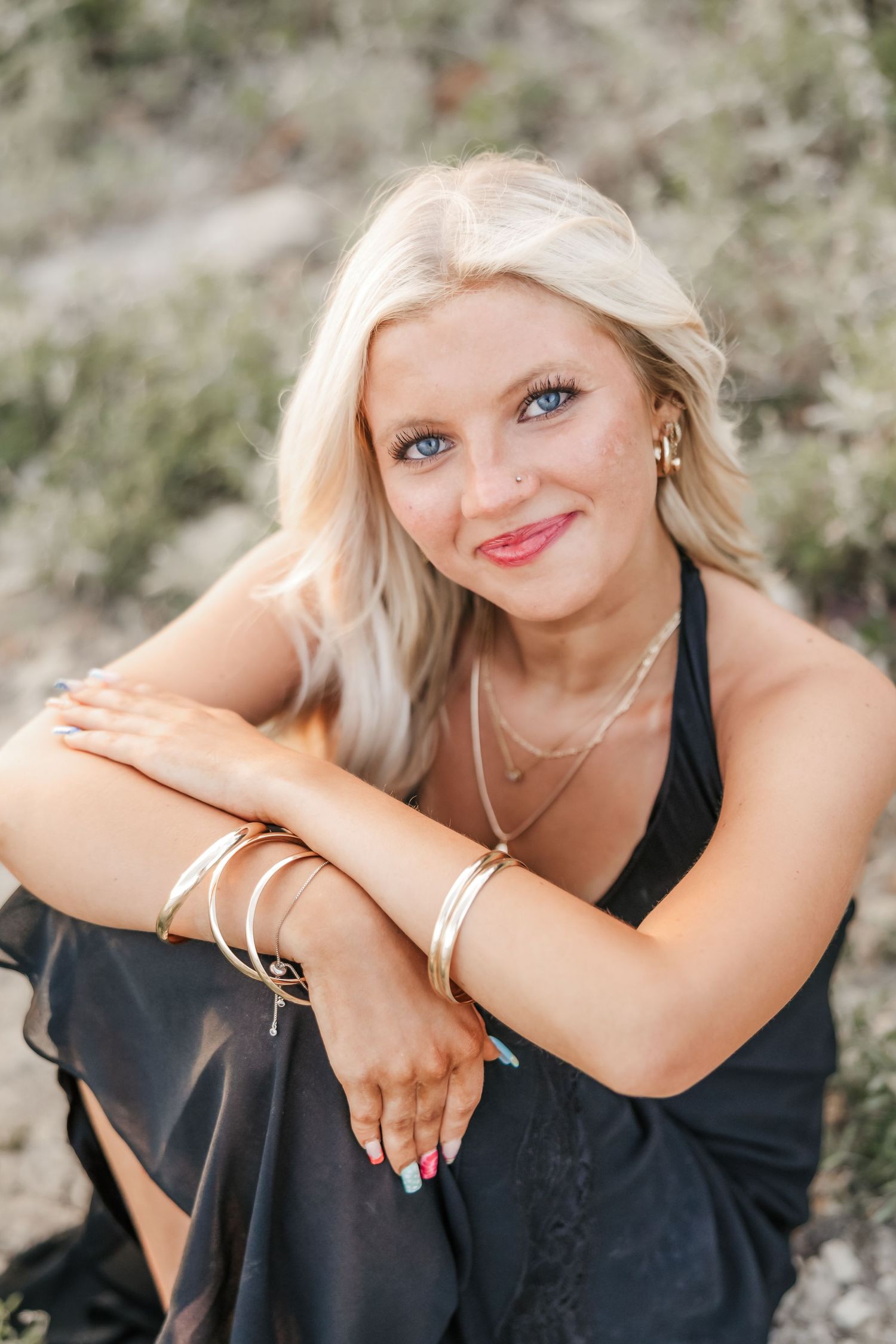Smiling blonde woman with blue eyes wearing a black dress and gold bracelets, seated outdoors in a natural setting.