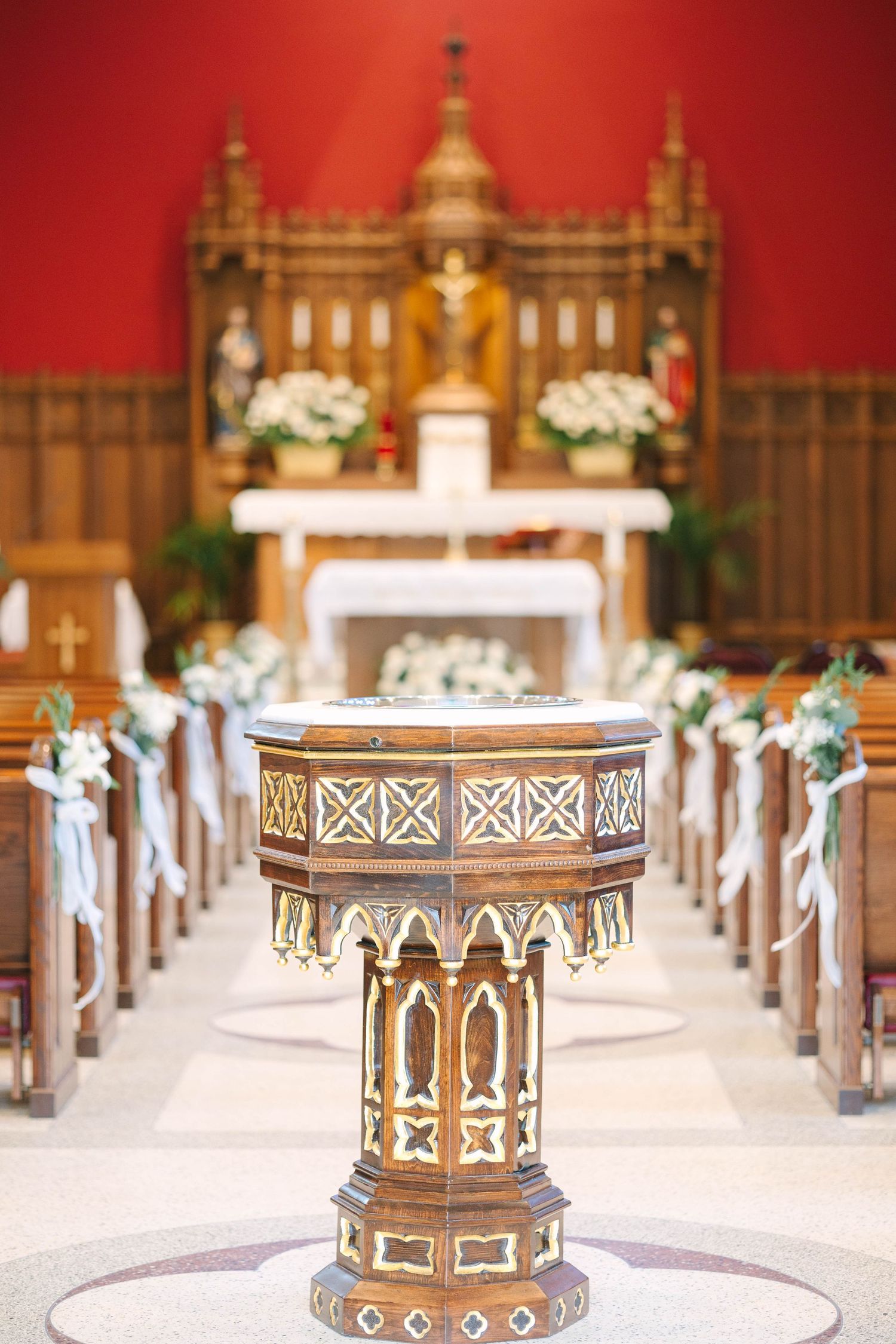 Ornate wooden baptismal font in a church aisle, with white floral pew decorations and a decorated altar under red walls.