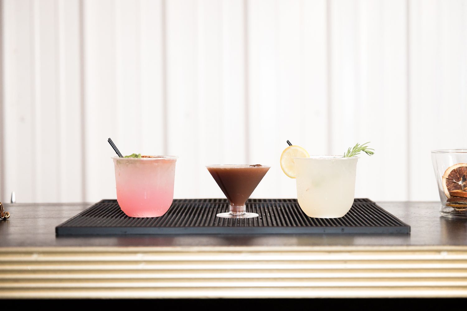 Three colorful cocktails in different glassware arranged on a black bar mat against a white background.