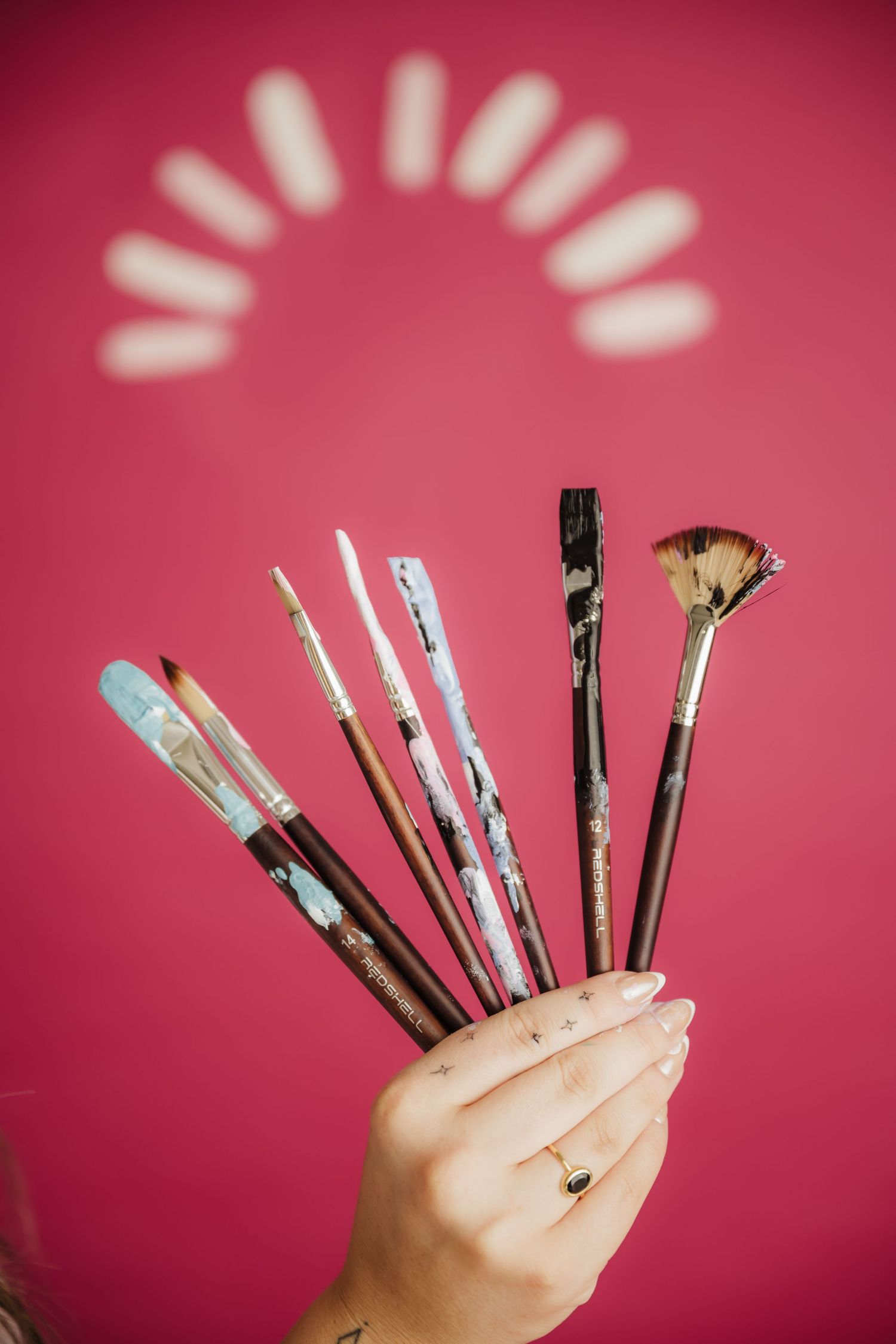 Hand holding a fan of paint brushes with colorful handles against a vibrant pink background with decorative arch.