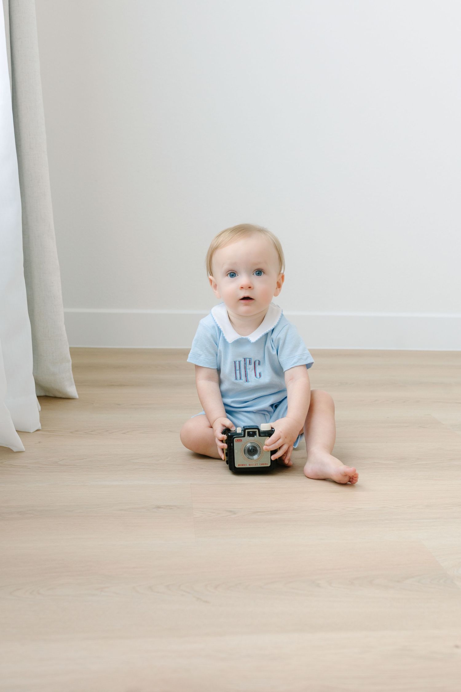 Adorable blue-eyed baby in light blue monogrammed romper sits on hardwood floor holding a vintage camera. Northlake, Tx in home photography