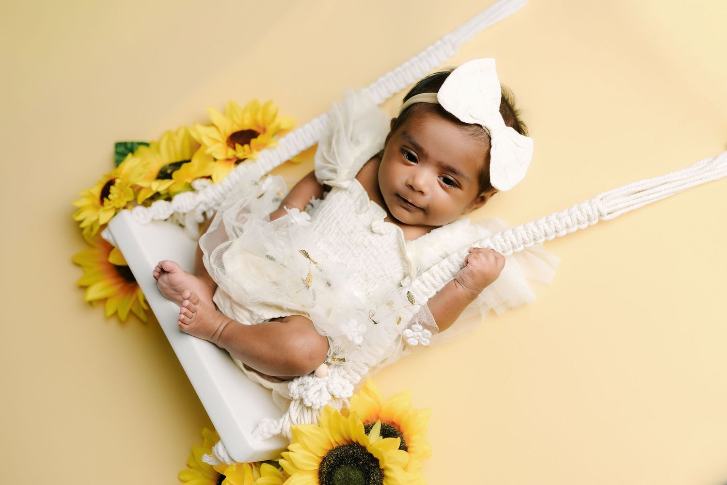 Smiling baby girl in white lace dress with bow headband lying in white frame surrounded by sunflowers on yellow background.