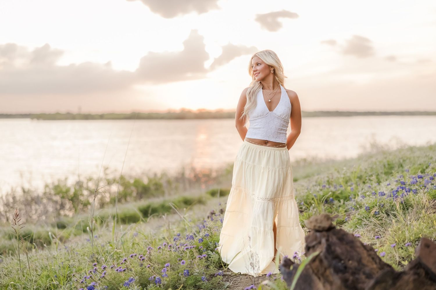 Blonde woman in white crop top and cream maxi skirt standing near a lake at sunset.