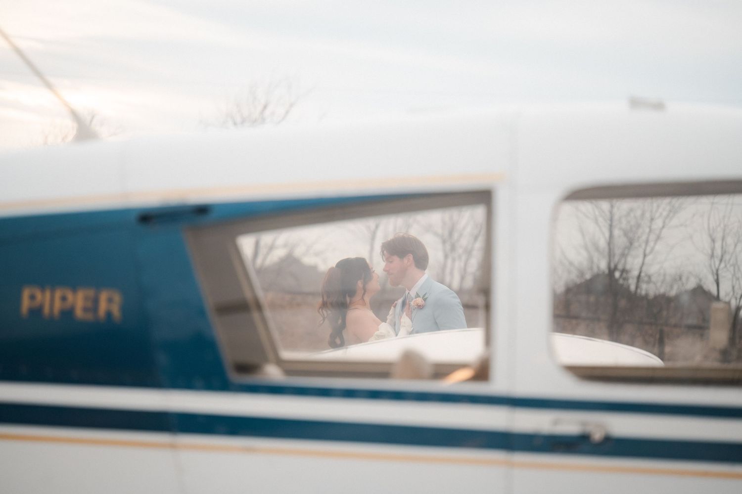 View through an airplane window showing a Piper aircraft with someone visible in the cockpit through reflective glass.