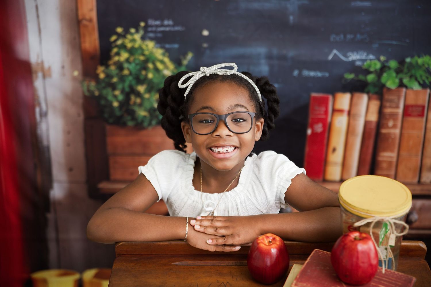 Smiling young girl with glasses sitting at a wooden desk with red apples and books in a classroom setting.