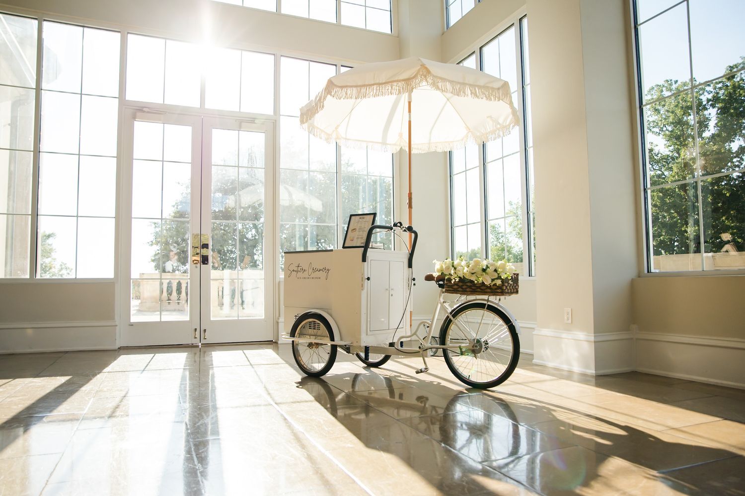 Bright sunlit venue with large windows and reflective wooden floors.