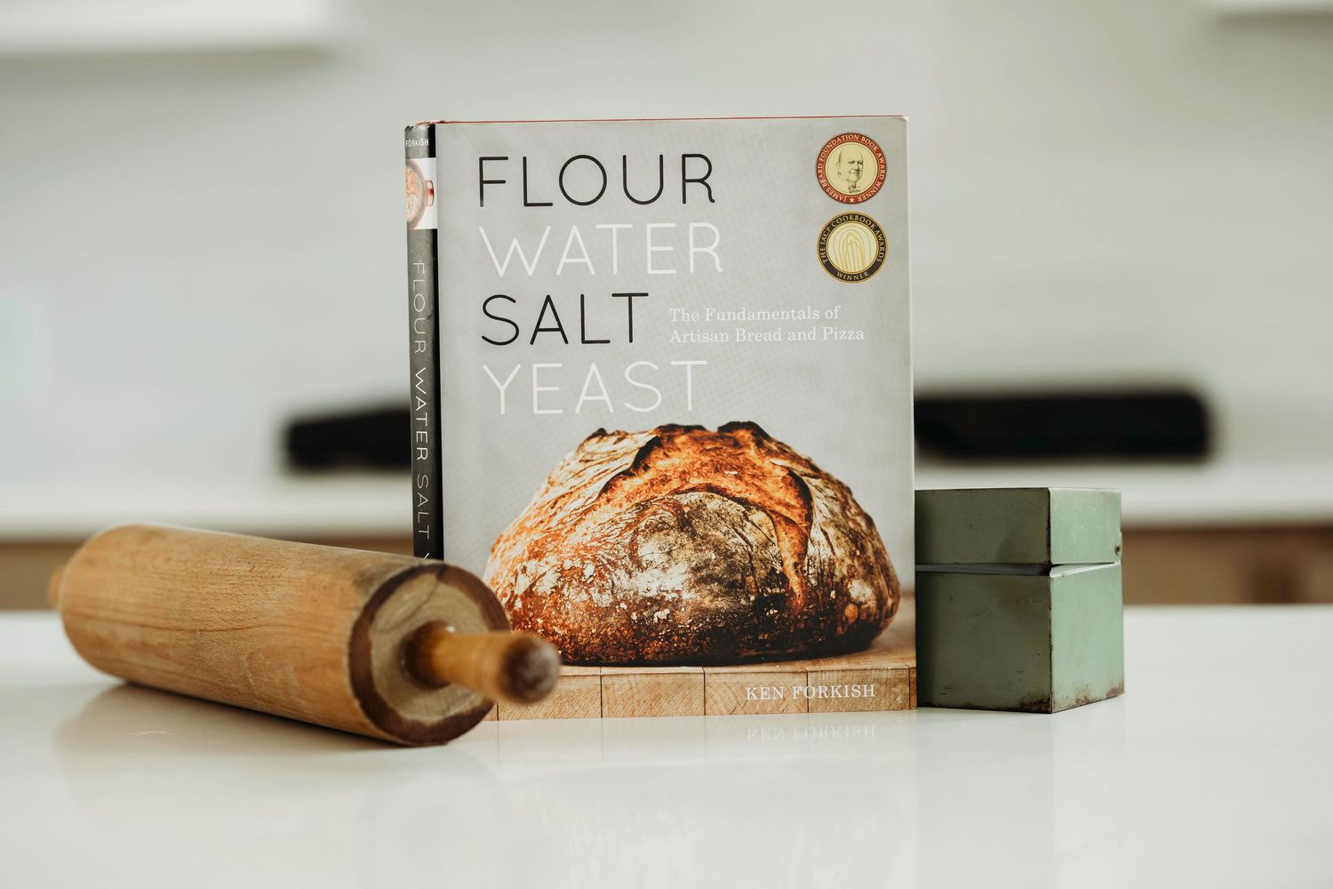 A wooden rolling pin and recipe book with 'Flour' and 'Salt' text sit on a white kitchen counter next to a green cube.