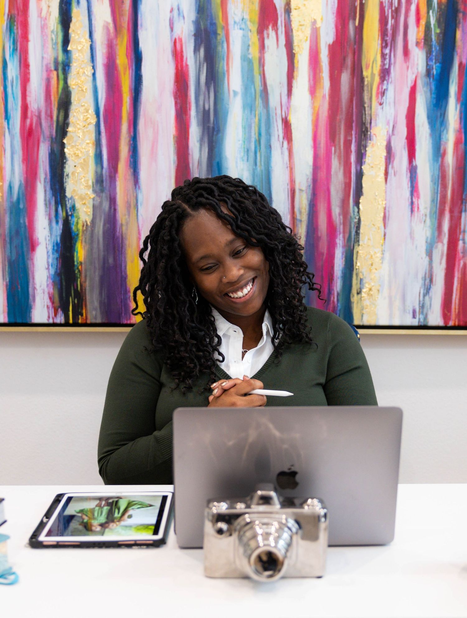 Professional wearing a dark blazer smiles while working on laptop with colorful abstract art displayed on wall behind.