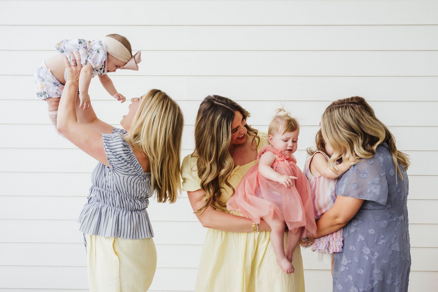 Three mothers in pastel dresses joyfully playing with their babies against a white shiplap wall.