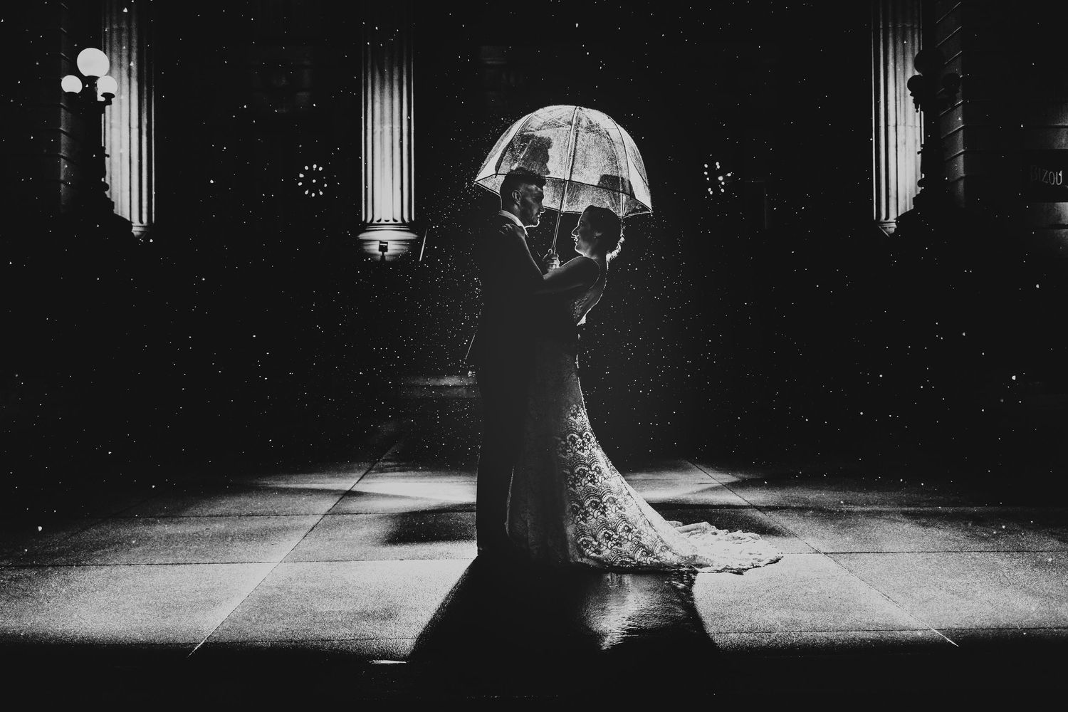 A silhouetted couple shares an intimate moment under an umbrella in a dramatic black and white photograph.