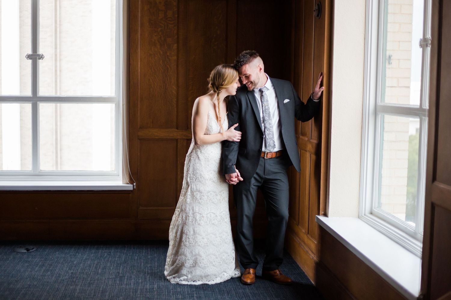 Elegant couple shares an intimate moment near sunlit windows in a rustic wood-paneled room.