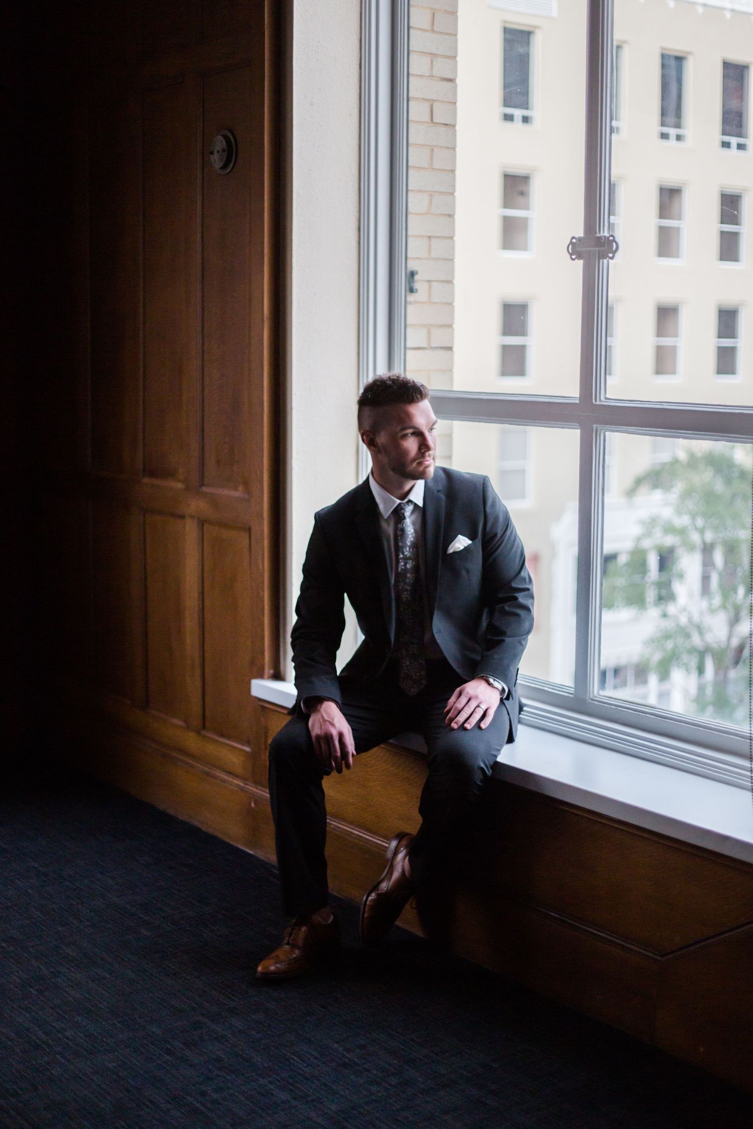 A groom  in a dark suit sits thoughtfully by a large window in a dimly lit room with wooden paneling, by Knot & Wing Photography