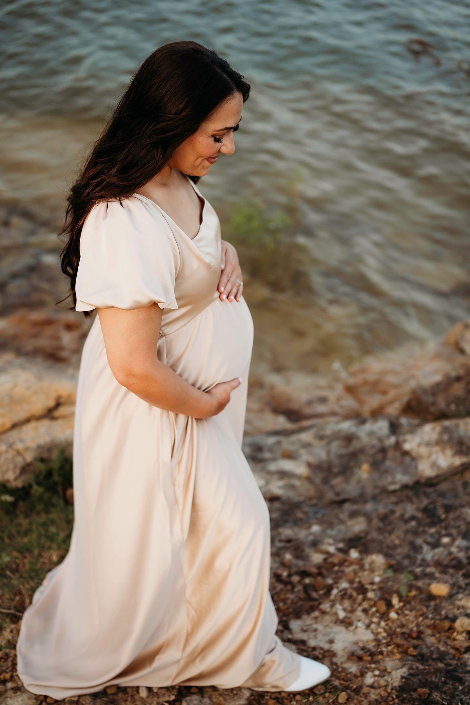 Expectant mother in a flowing white dress poses near the water's edge during golden hour maternity photoshoot.