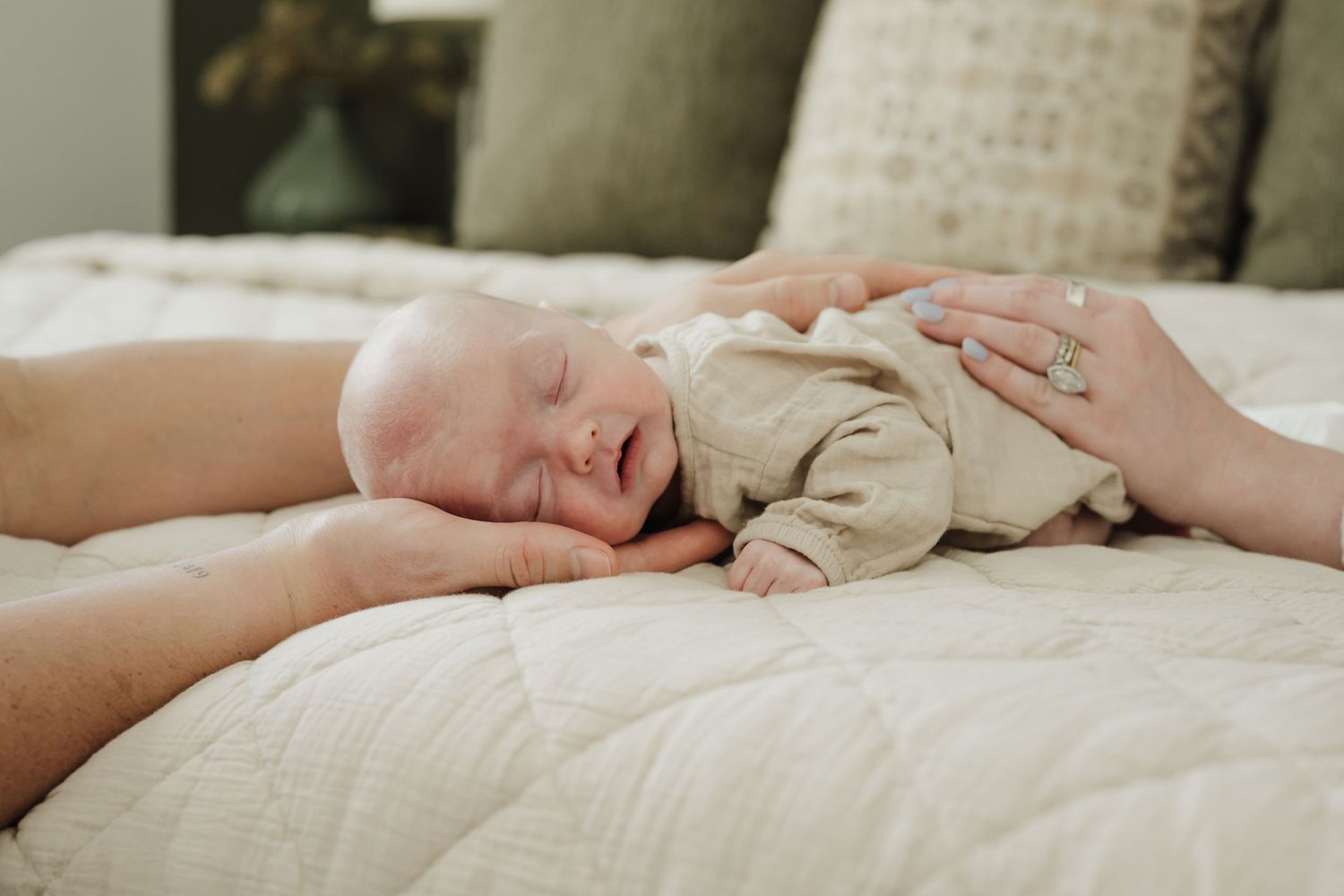 A sleeping newborn baby wearing a beige onesie rests peacefully on white bedding while being gently touched.