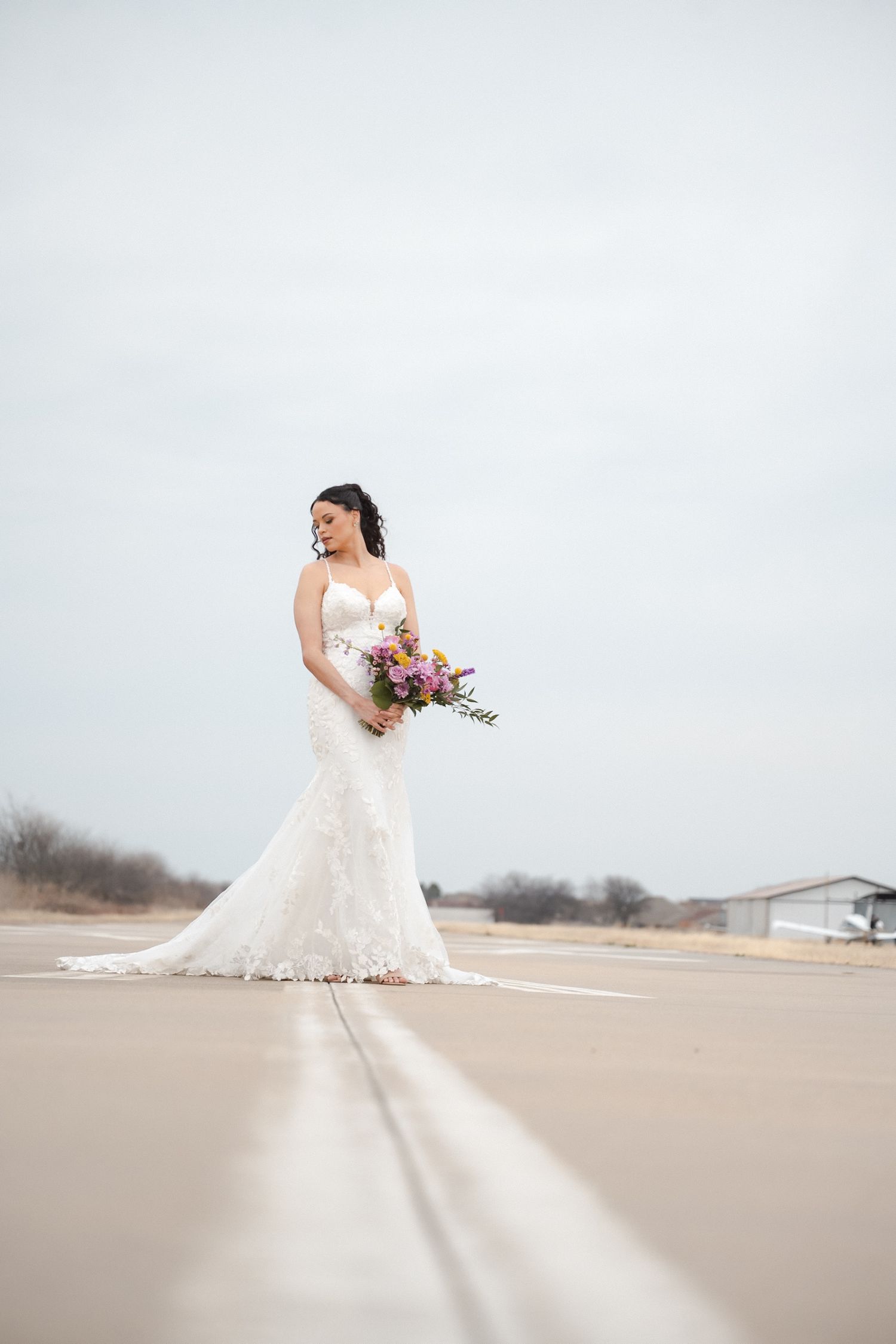 A bride in a flowing white lace dress holds a vibrant bouquet while posing on a cloudy winter day.