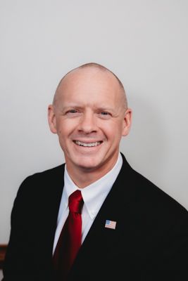 Professional headshot of someone in a black suit and red tie against a light gray background.