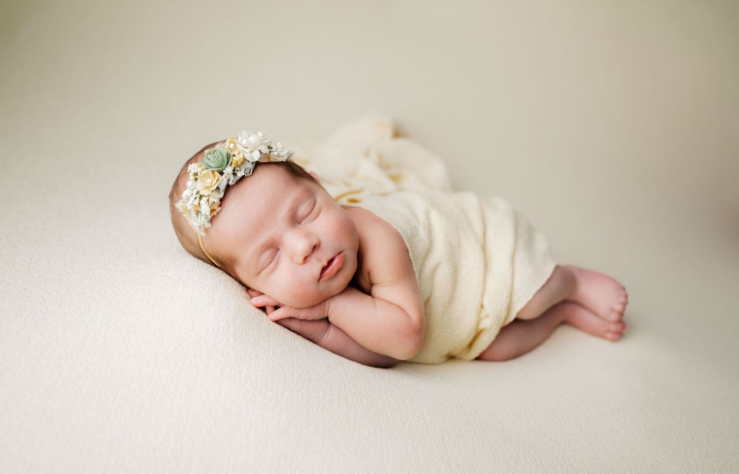 Peaceful newborn baby sleeping wrapped in cream fabric with floral headband on soft white background.