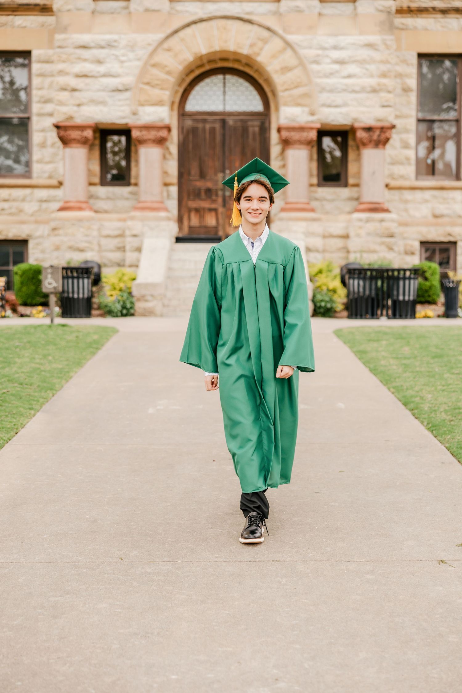 Smiling graduate in green cap and gown walking on pathway in front of stone building.