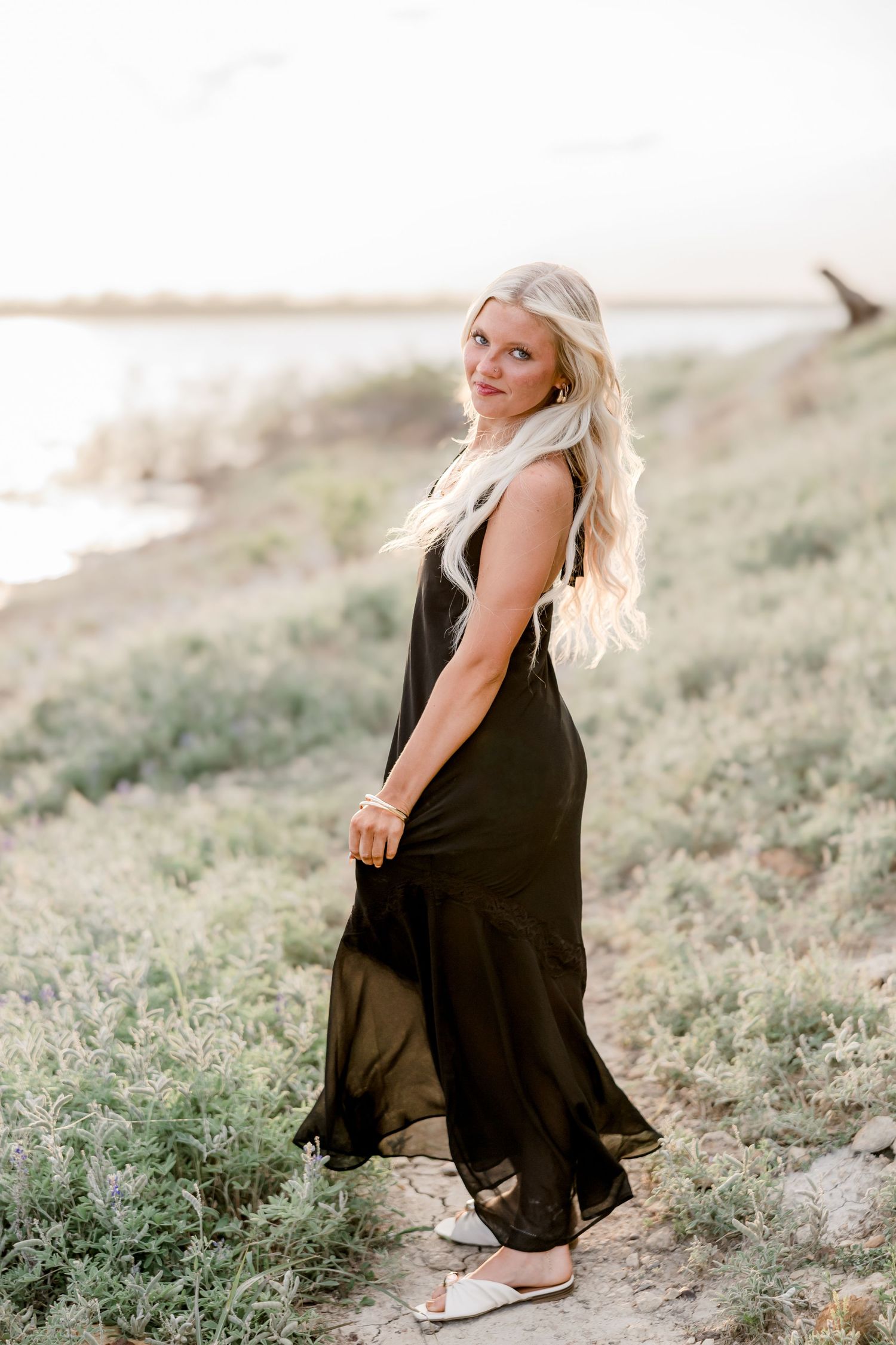 Blonde woman in black maxi dress posing on a grassy hillside near water at golden hour.