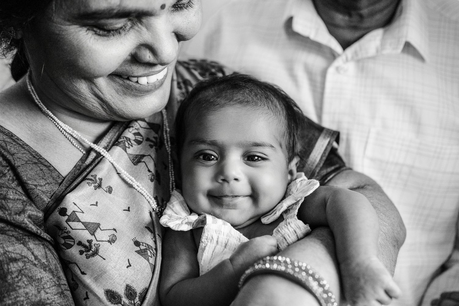 Black and white photo of a smiling Indian woman holding a happy baby, with a man partially visible in the background.