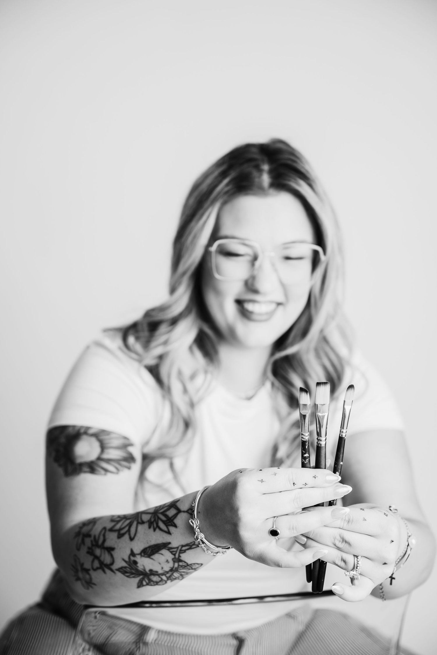 Smiling woman with tattoos holding paint brushes, wearing glasses and a white t-shirt, black and white photo.