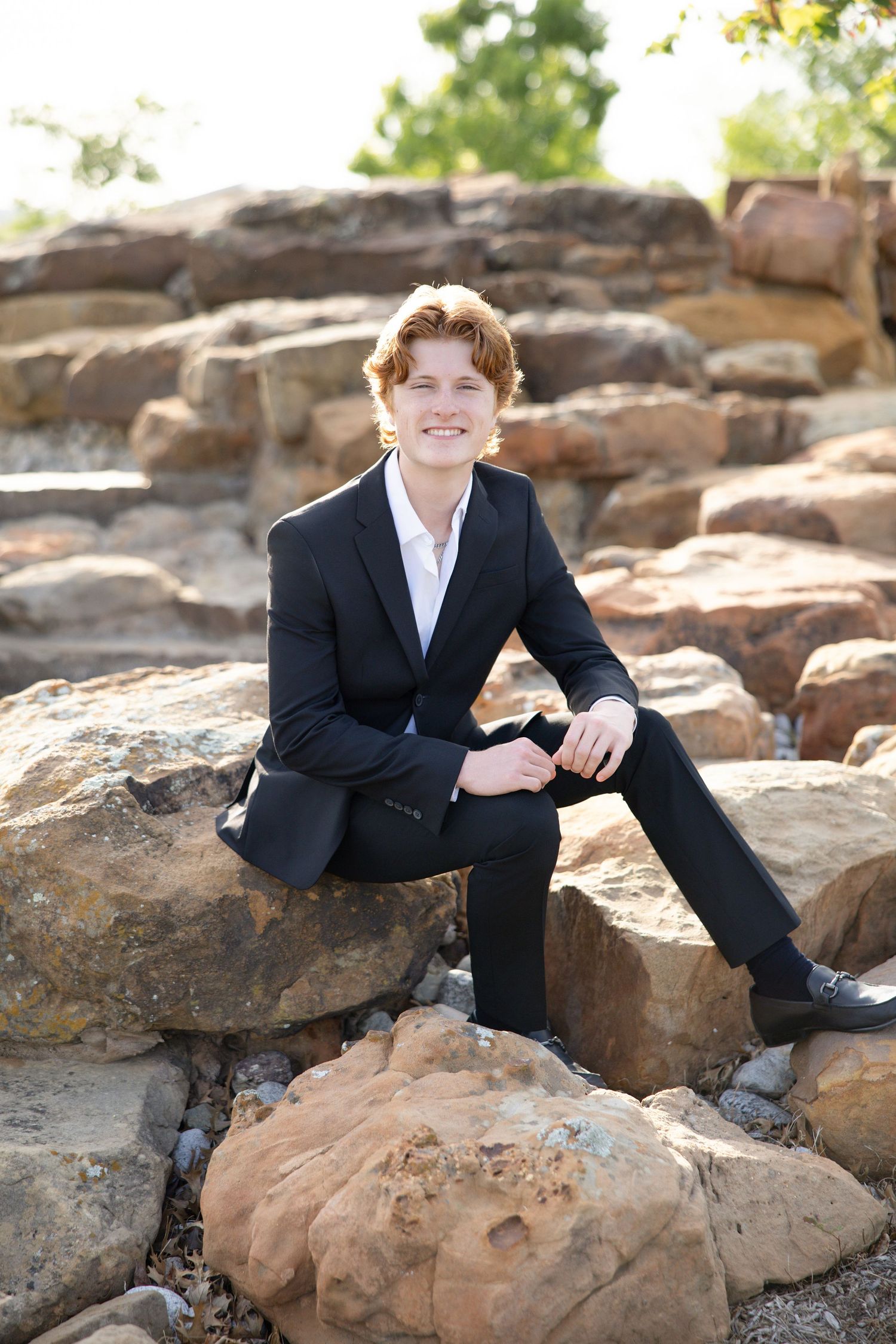 Young man with red hair in black suit sitting on rocks outdoors, smiling confidently for a portrait photo.