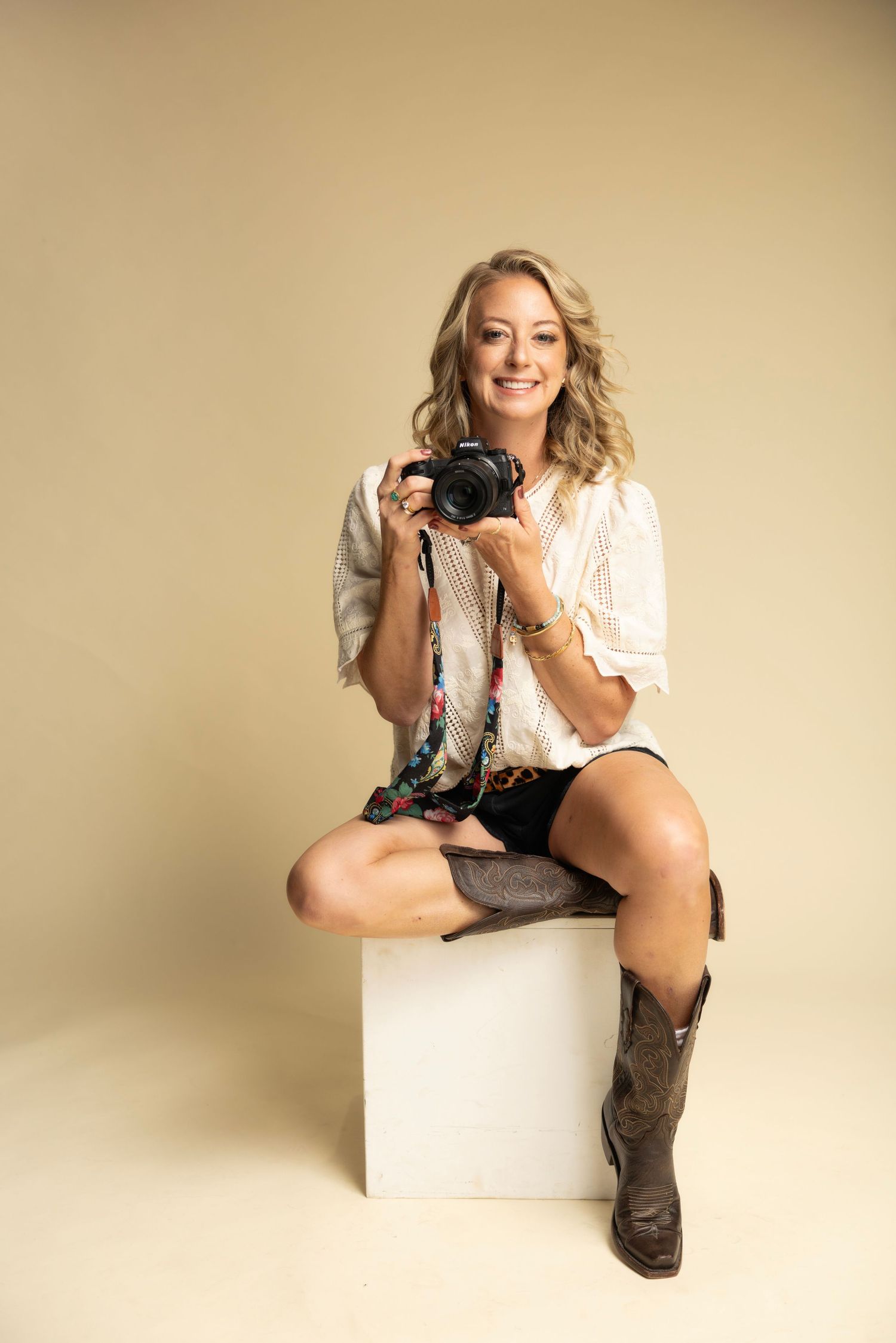Professional photographer wearing boots sits casually on white cube while holding camera in studio setting.