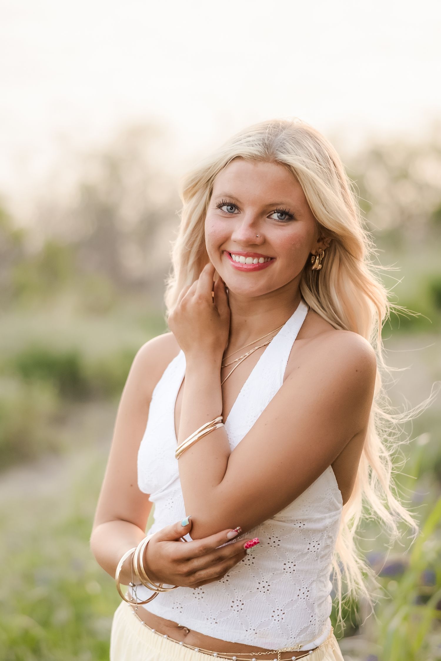 Smiling blonde woman in white halter top with gold jewelry posing outdoors in a sunlit field.