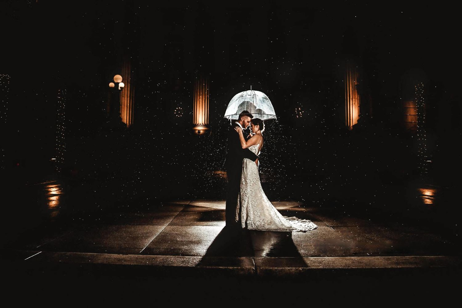 A couple dances in an elegant dark venue with dramatic backlighting creating a romantic silhouette effect.