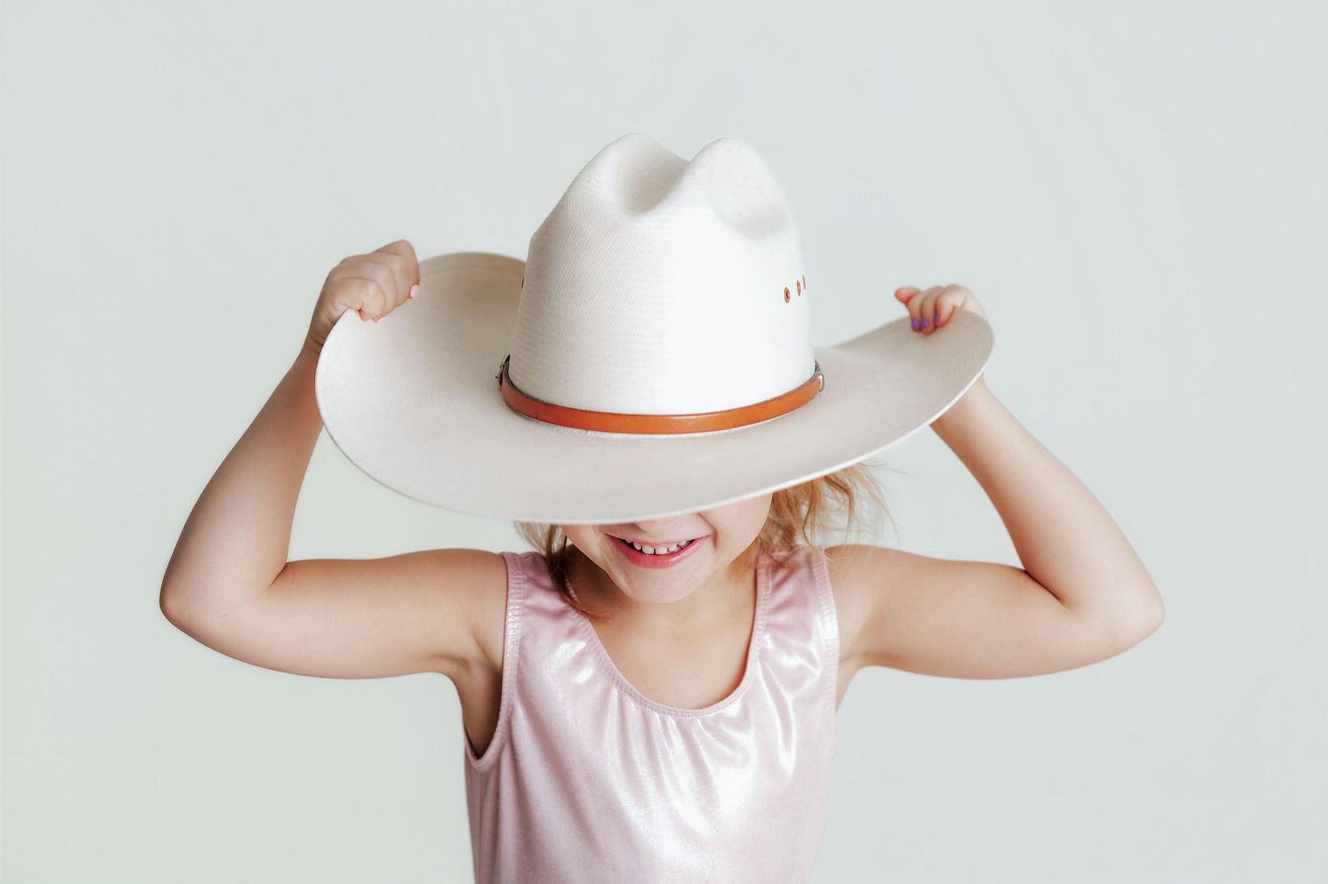 A person in a white sunhat and pink dress poses playfully while smiling against a light background.