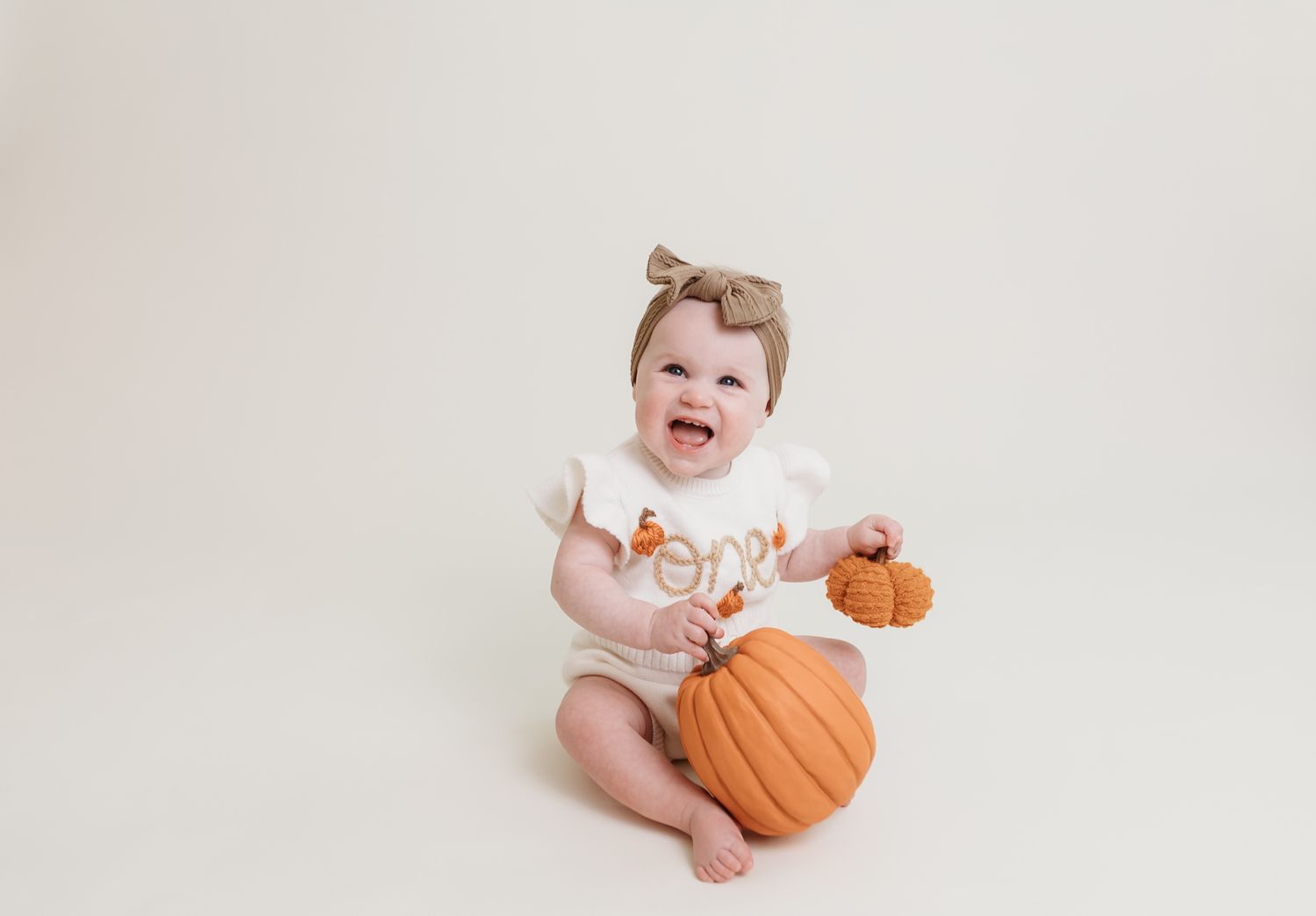 A baby in a white onesie sits next to a large orange pumpkin against a light colored background.