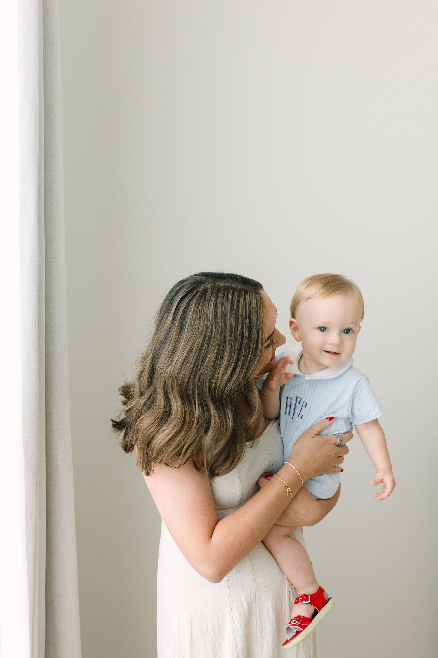 A mother holds her smiling baby dressed in a light blue onesie and red shoes against a white background.