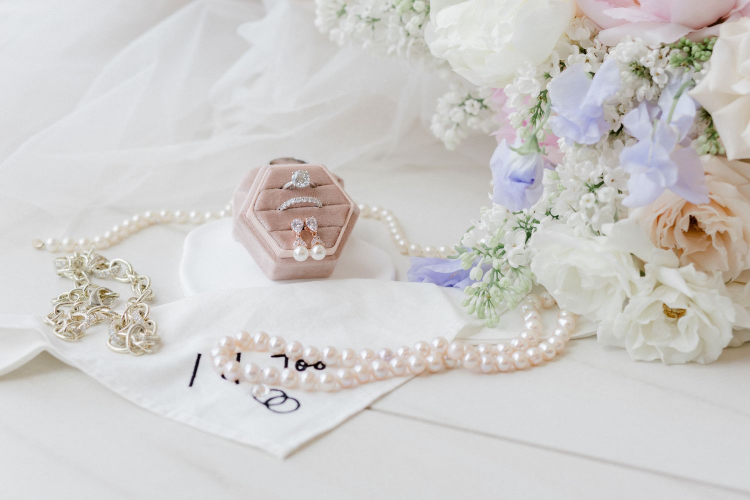Elegant wedding flat lay with pearl necklace, pink velvet ring box, earrings, and pastel floral bouquet on white background.