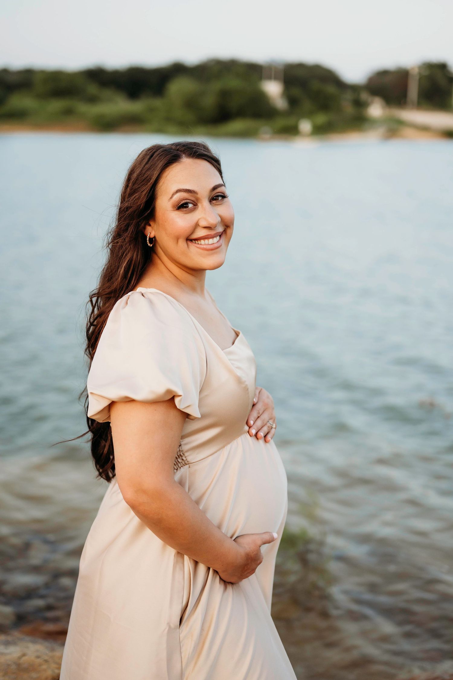 Pregnant woman in light-colored dress smiles joyfully by the lake during sunset maternity session.