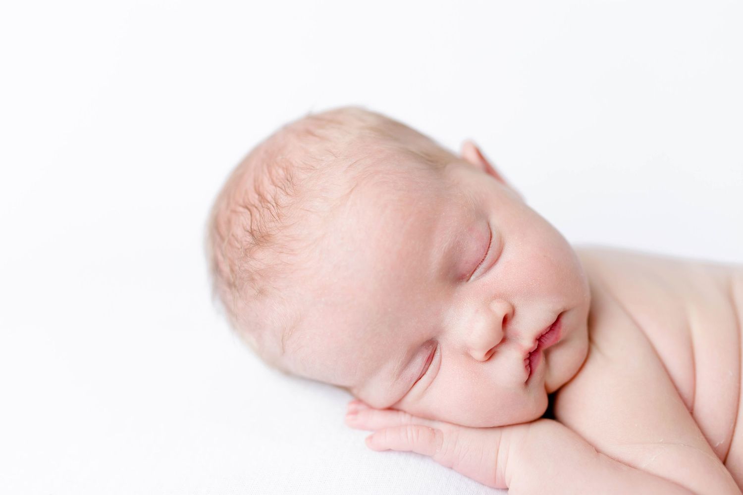 A sleeping newborn baby lies peacefully on a soft white blanket in a minimalist portrait setting.