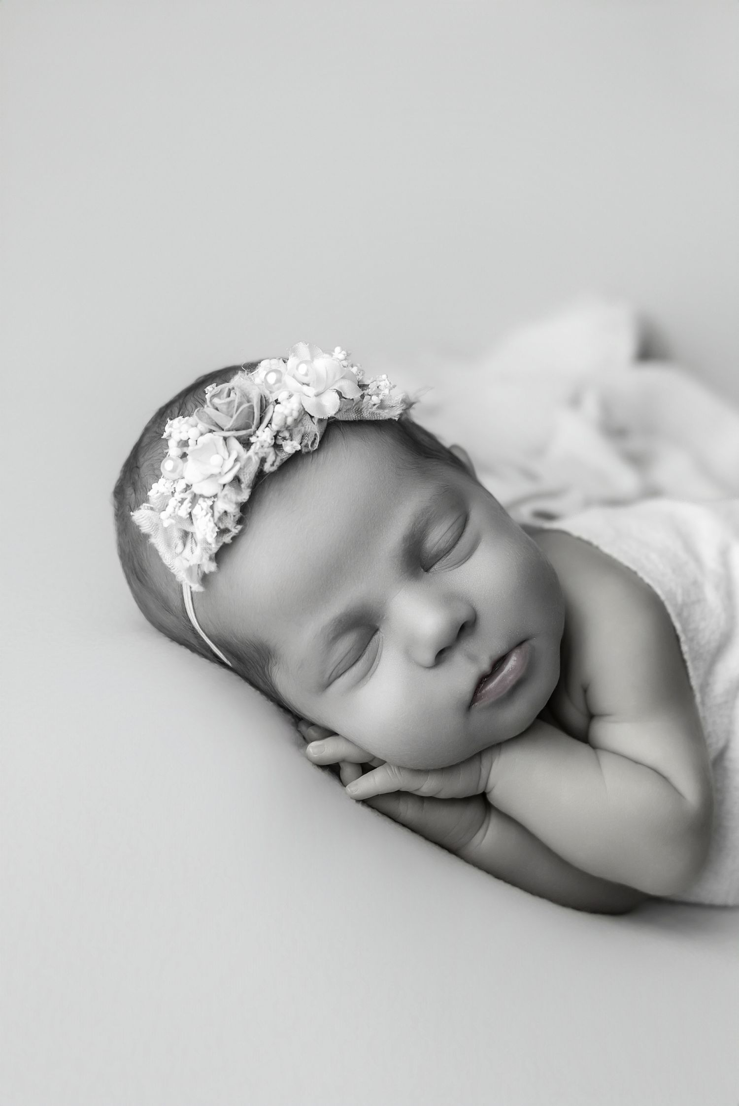 Black and white newborn photo of a sleeping baby girl wearing a floral headband, resting her chin on her hands.