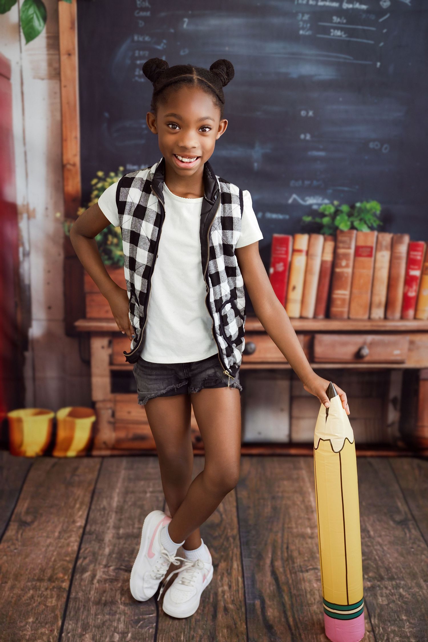 Smiling young girl in plaid vest holding a giant pencil prop, standing in front of a chalkboard classroom backdrop.