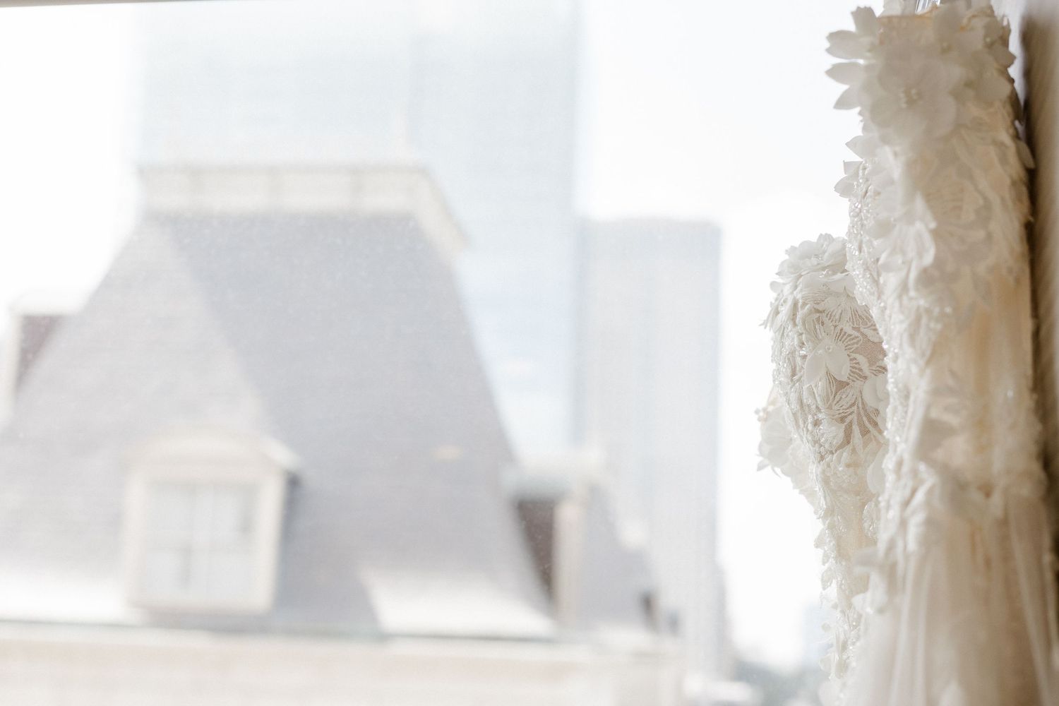 White lace wedding dress hanging near a window with blurred rooftop view in the background.