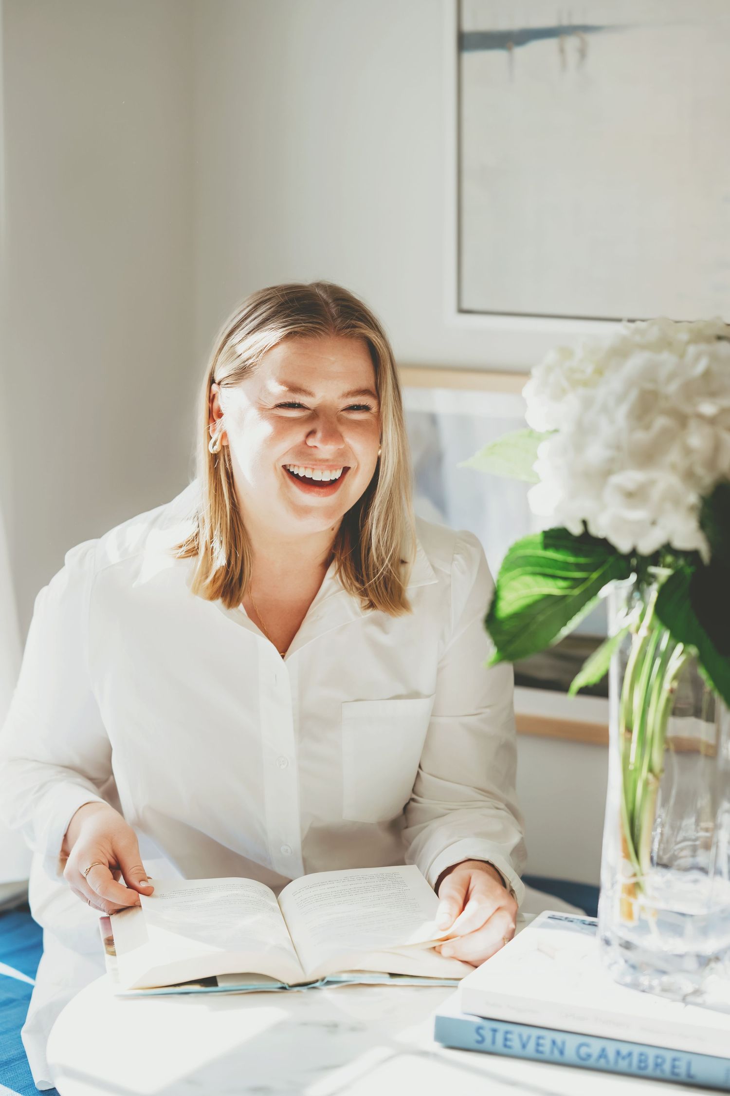 Woman in white shirt laughs joyfully while reading a book in a bright, cozy interior setting.
