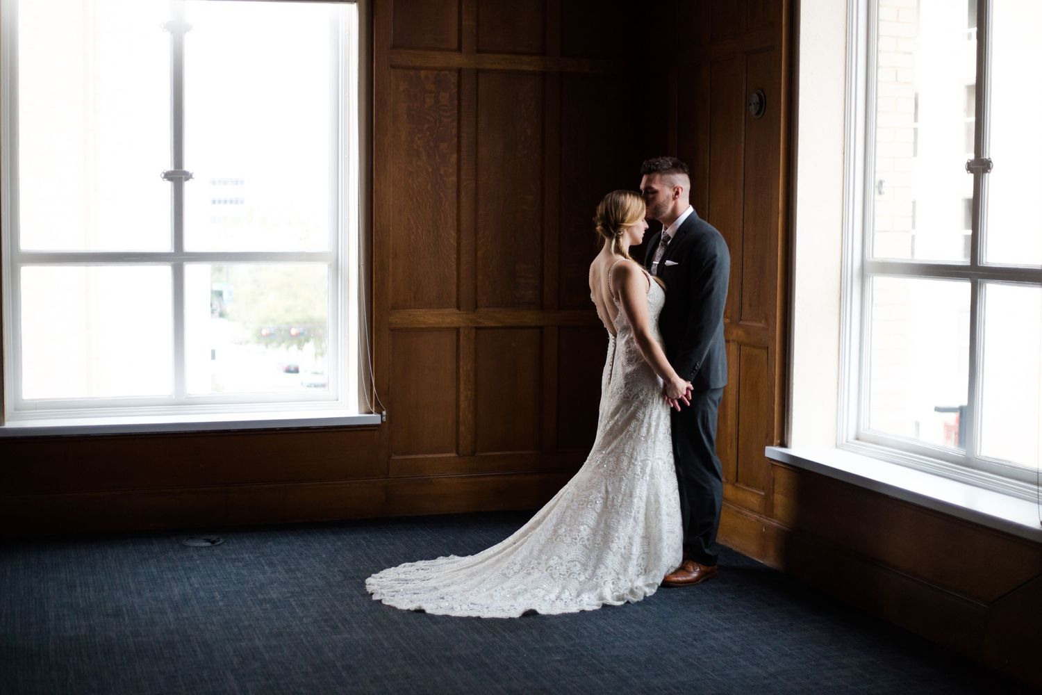 Bride and groom share an intimate moment in a sunlit room with dark wood paneled walls.